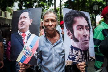 | A man in Bolívar Square with Simón Bolívar and Chávez portraits and a copy of 99s Venezuelan Constitution | MR Online